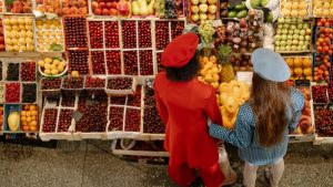 Two women shopping at a colorful fruit market, viewed from above, showcasing a variety of fresh fruits.