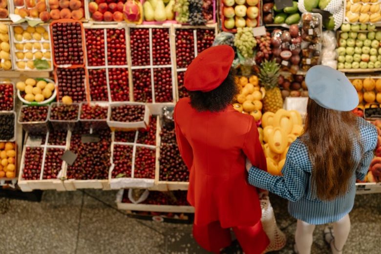Two women shopping at a colorful fruit market, viewed from above, showcasing a variety of fresh fruits.
